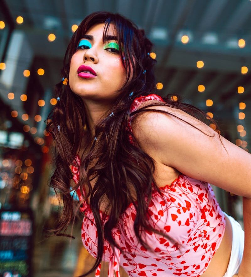 A fashionable woman with vibrant makeup poses under decorative string lights at night.