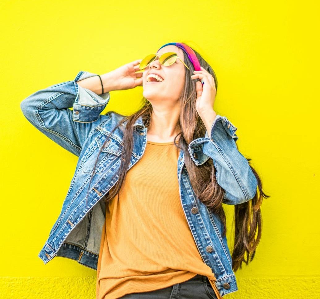 Smiling woman in sunglasses stands against vibrant yellow wall, radiating happiness.