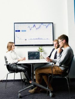 Three business professionals in a meeting room discussing financial charts displayed on a screen.
